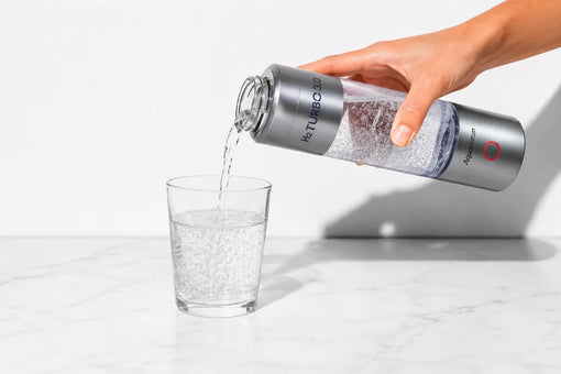 Hand gießt molekulares Wasserstoffwasser aus der silbernen H2 Turbo 3.0 Flasche in ein Glas auf einer Marmorplatte
